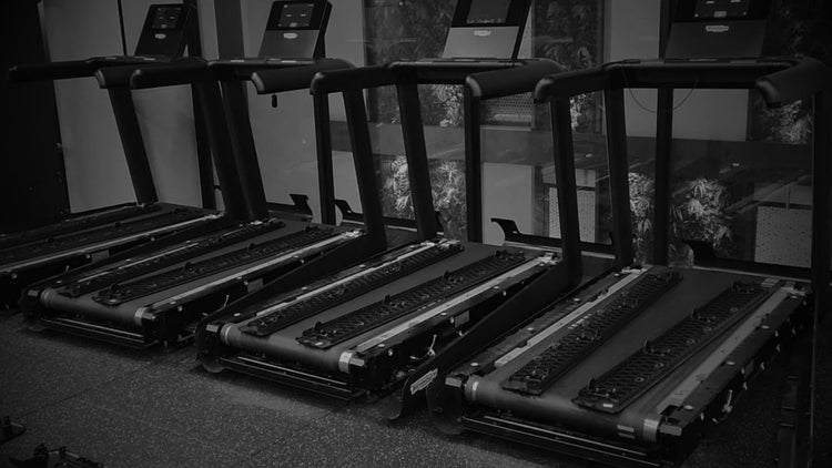 A row of black treadmills lined up in a gym setting, captured in a dark, high-contrast black and white photograph.